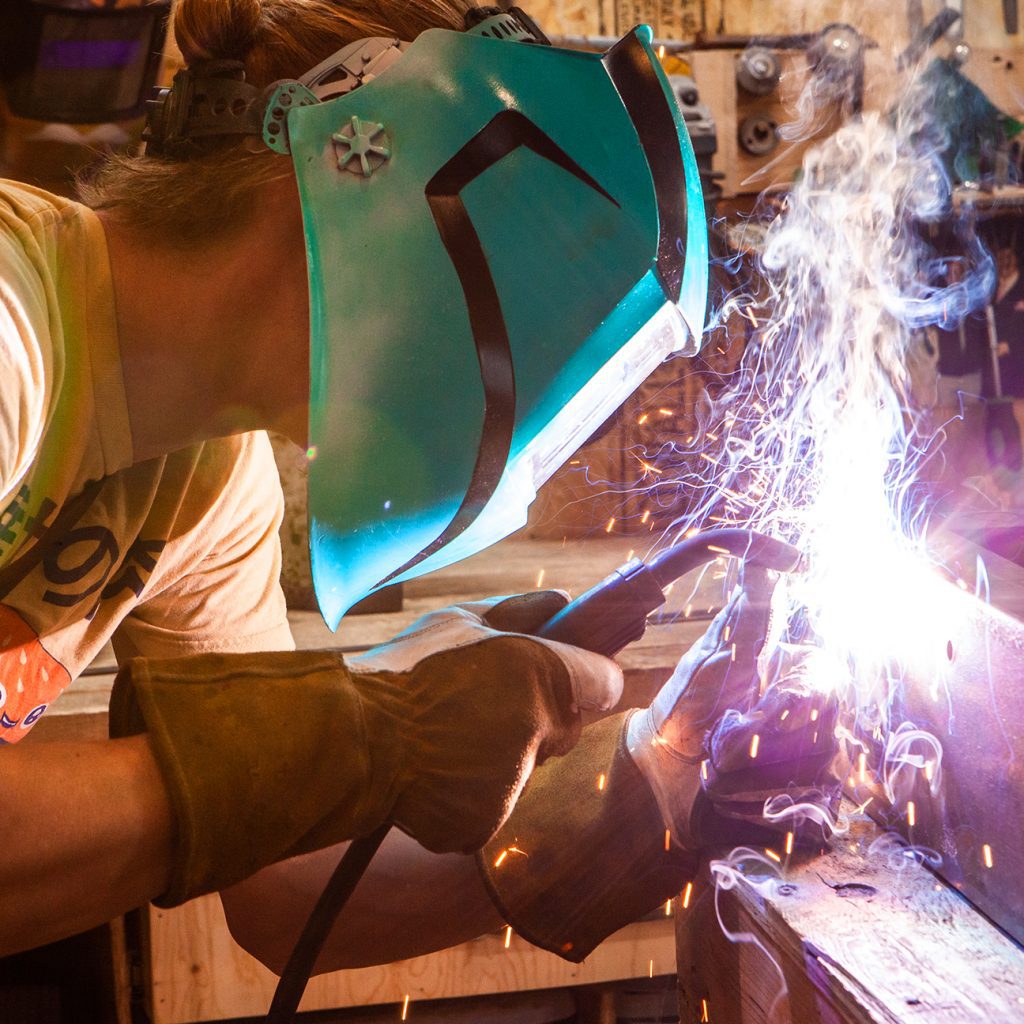 A closeup and side profile of a metalworker using a gas metal arc welding machine to join two pieces of steel. Skilled worker operates welding machine.
